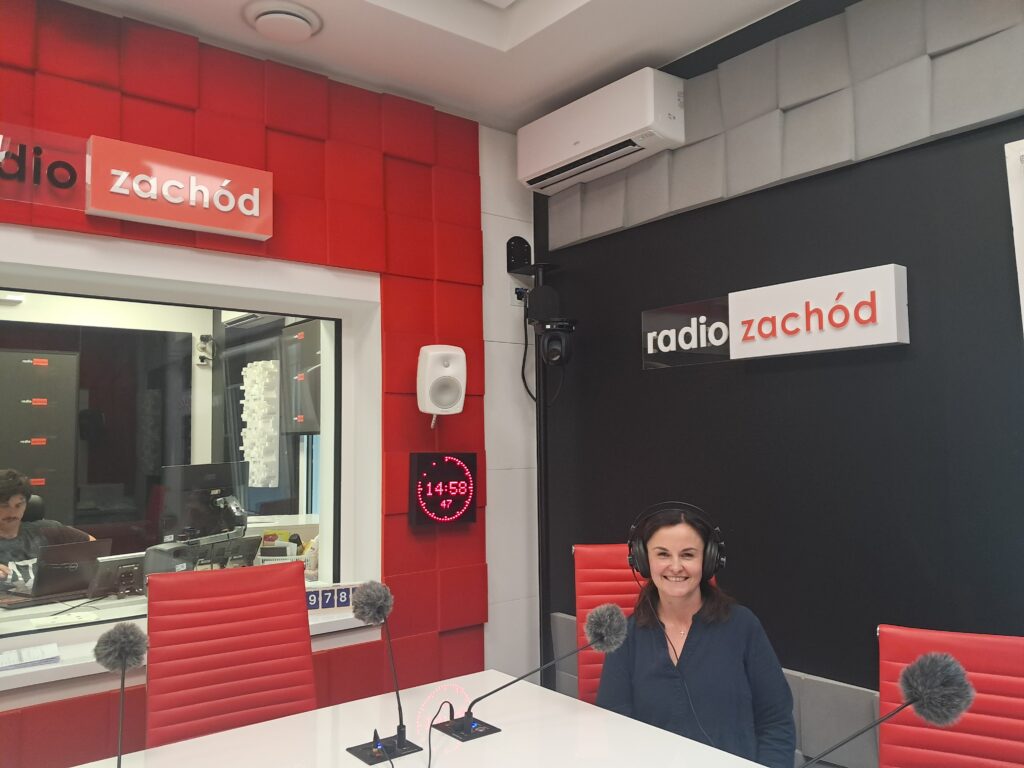 Kobieta uśmiecha się siedząc przy biurku w studiu radiowym z mikrofonami, otoczona czerwonymi i czarnymi panelami akustycznymi oraz cyfrowym zegarem pokazującym 14:58. Atmosfera jest profesjonalna, spokojna i przyjazna.
