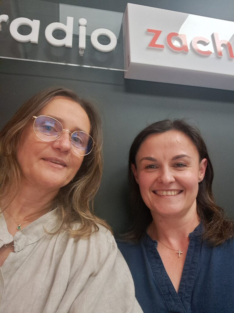Dwie kobiety, jedna w okularach i jasnej koszuli, druga w niebieskim topie, obie uśmiechają się do kamery. Tło stanowi szara ściana z białym i czerwonym napisem 'Zach Radio'